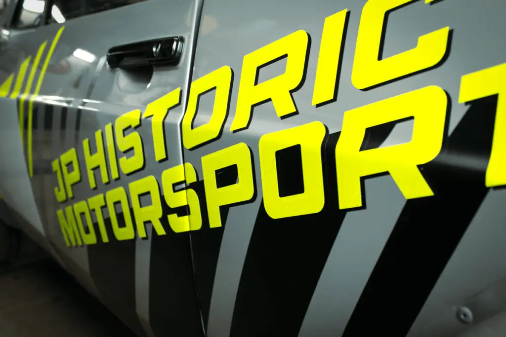 Logo of JP Historic Motorsport on the side of the rally car. In Fluo Yellow and black outlines.