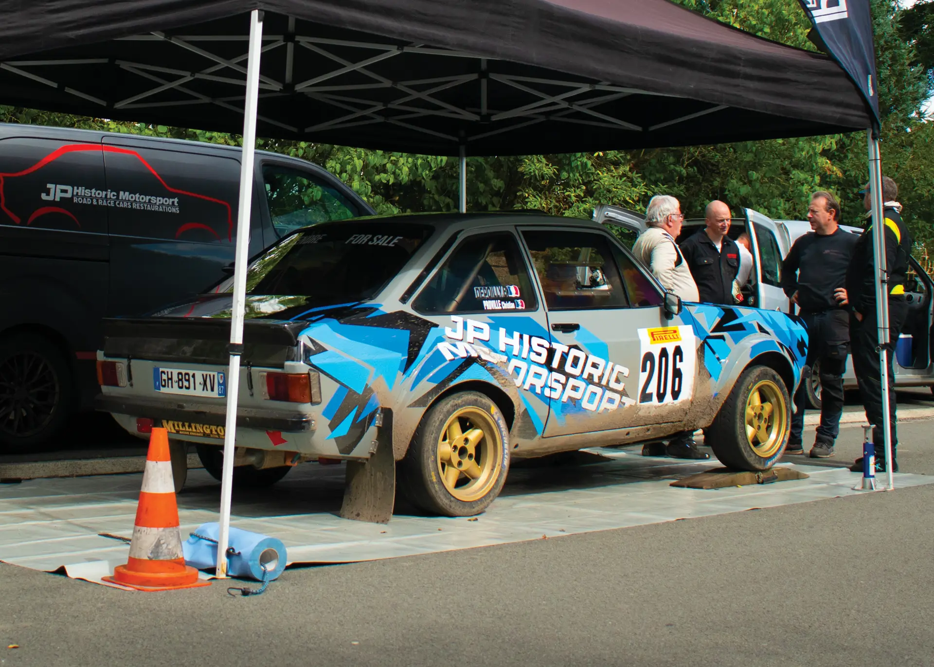 A classic rally car, adorned with blue and black livery and marked with the text "JP Historic Motorsport," sits under a tent on a paved surface. The car displays the number "206" and wears rally-style mud flaps and yellow wheels. A small "For Sale" sign is visible on the rear windshield. Nearby, several men are engaged in conversation, with a branded van in the background and various rally equipment like a traffic cone and roll of blue tape placed around the vehicle. The environment suggests a motorsport or rally event.